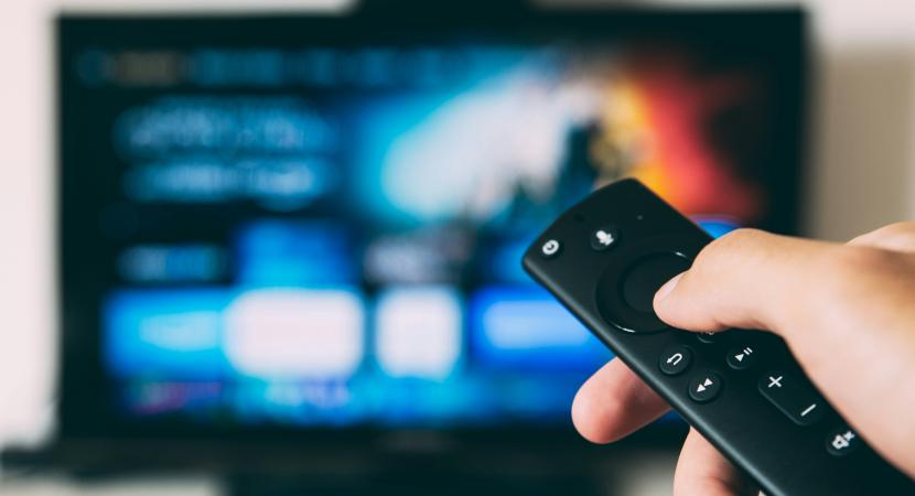 Photograph of a TV remote pointed at a television screen in the background; the screen is blurry but the shapes of online streaming applications is visible